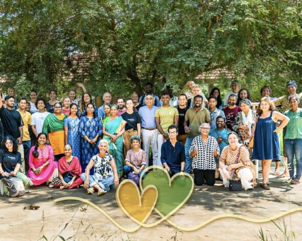 Diverse group of people proudly connected at Auroville International USA event under a large tree.
