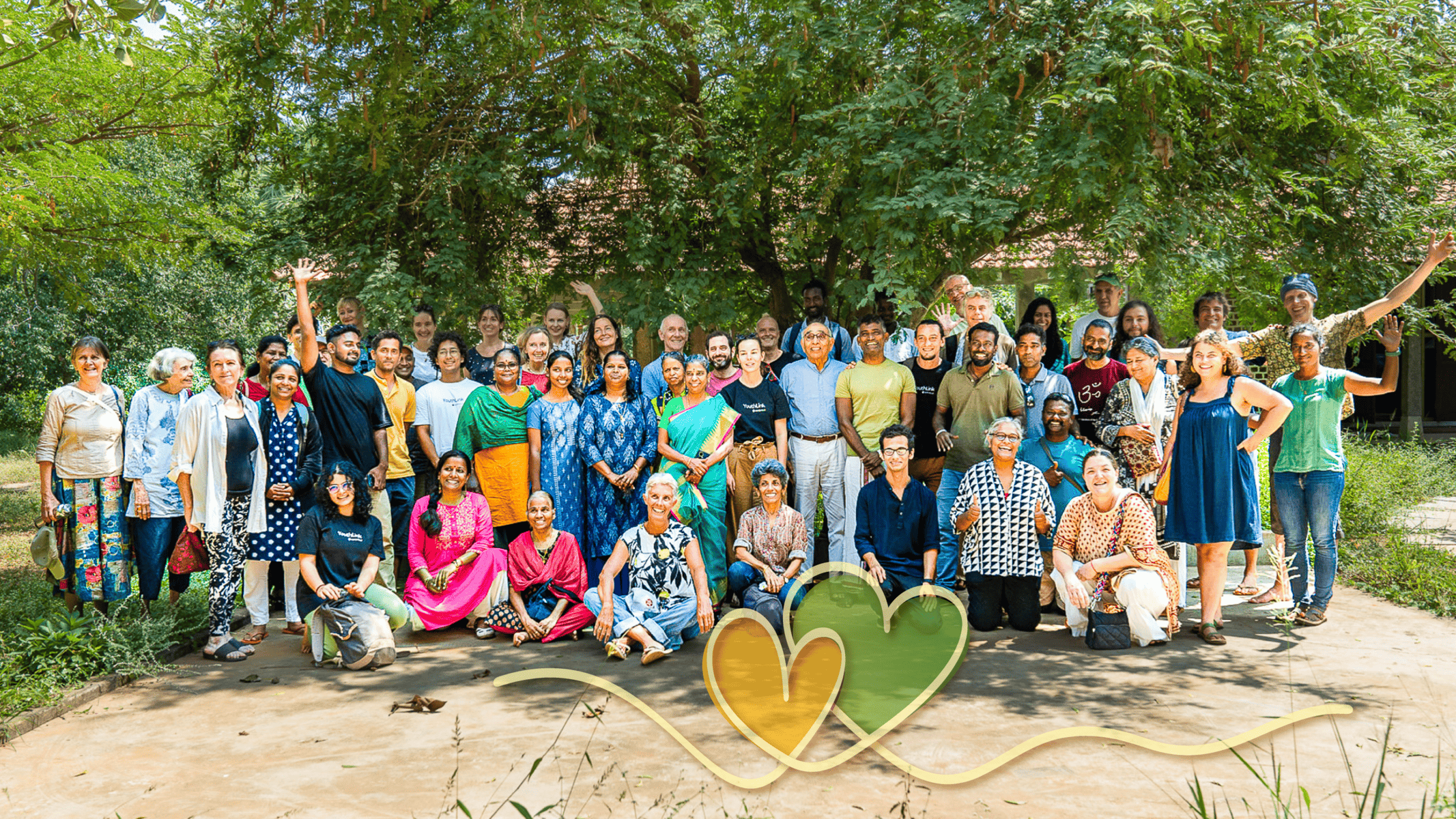 Diverse group of people celebrating at Auroville International USA event under a large tree.